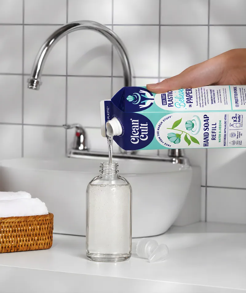 Pouring a hand soap refill carton into an aluminum bottle at a bathroom sink